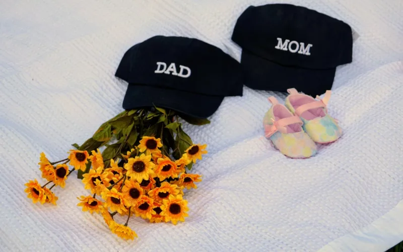 Baby shoes with “Mom” and “Dad” caps beside yellow flowers.