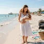 Mother holding a newborn baby while standing on a sunny beach near the shoreline with a stroller and beach bag nearby.