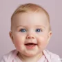 Smiling baby with bright blue eyes and rosy cheeks, wearing a light pink shirt.