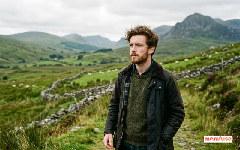 young irish/scottish man with light beard, thoughtful expression, green countryside background.