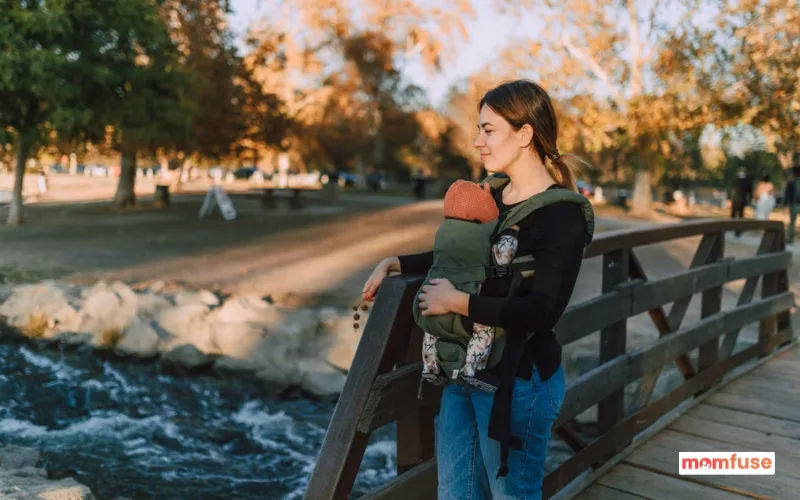 Mother carrying her baby in a front baby carrier while standing on a wooden bridge in a park.