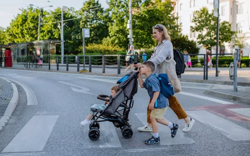 Woman walking across a city street with two children, pushing a stroller during a trip.