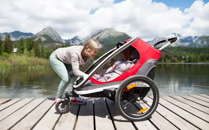 Woman adjusting a red jogging stroller with a baby on a wooden dock by a mountain lake.