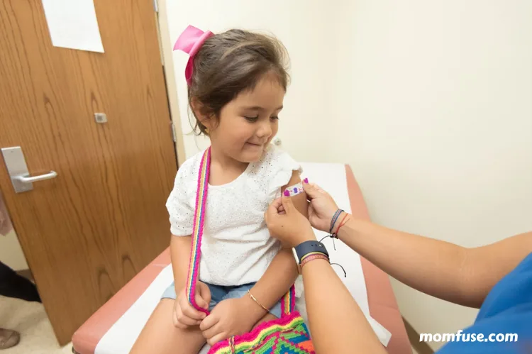 A young girl sits on a medical exam table smiling as a healthcare worker places a bandage on her arm after vaccination.