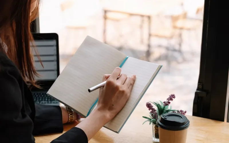 Woman writing notes in a notebook beside a laptop and coffee cup.