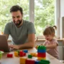 A smiling stay-at-home parent working on a laptop at a kitchen table while watching their child play.