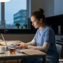A modern mother in scrubs studying on a laptop at a kitchen table late evening.
