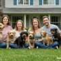 A cheerful family sitting on the grass in front of a cozy suburban home, surrounded by various dog breeds.