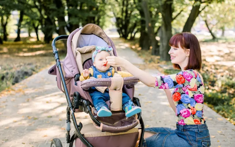 Woman feeding a baby sitting in a stroller on a park path.