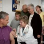A doctor administers a vaccine to a man while several people, including children and seniors.