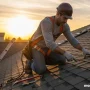 A skilled roofer wearing safety gear stands on a residential roof at golden hour inspecting shingles.