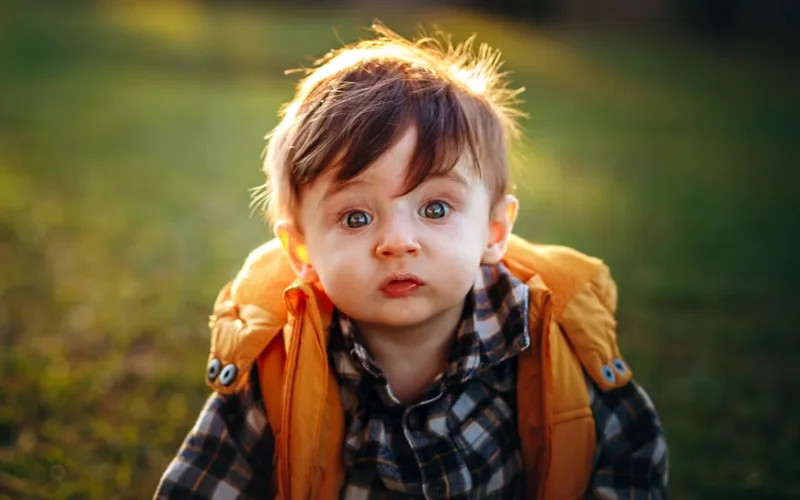 Cute baby boy wearing a yellow jacket outdoors in a green natural setting.