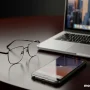 A clean, modern image of stylish eyeglasses placed next to a laptop and smartphone on a dark background.