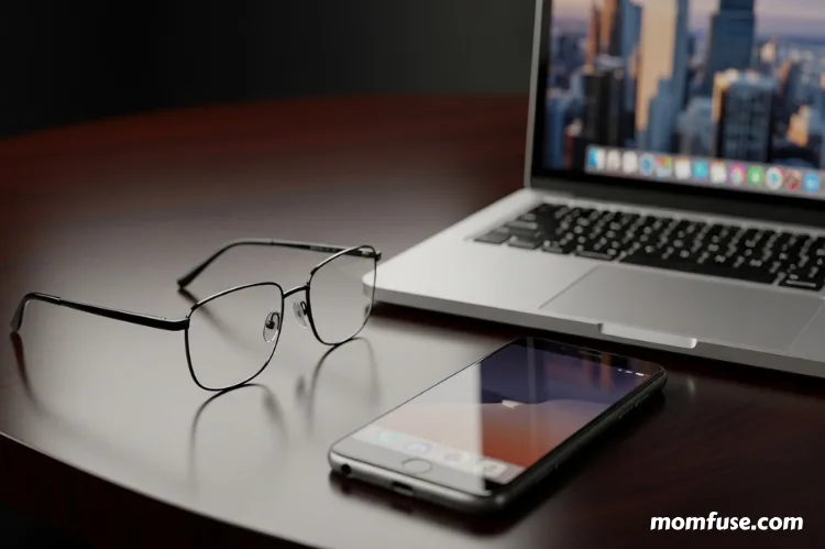 A clean, modern image of stylish eyeglasses placed next to a laptop and smartphone on a dark background.