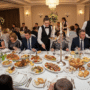 A wide banquet table filled with gourmet dishes, elegant tableware, and guests enjoying food.