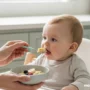 A warm, comforting scene of a parent gently feeding a baby from a small bowl of wholesome food.