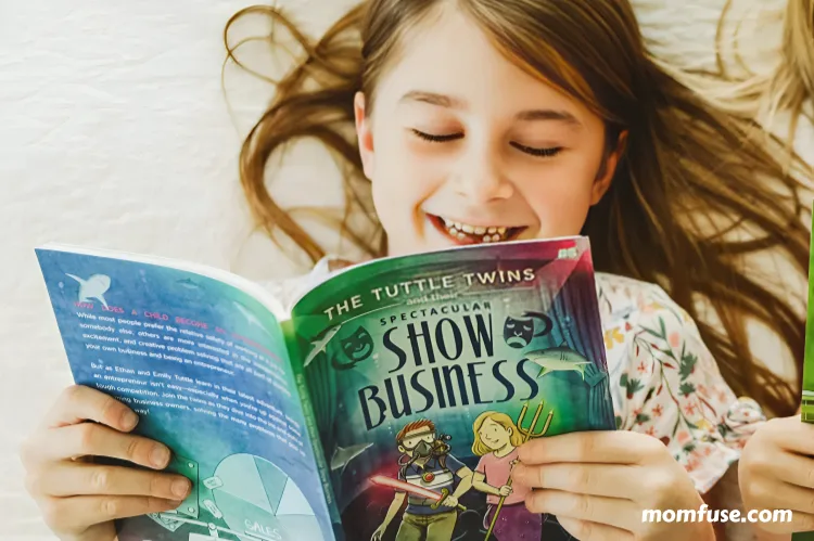A smiling young girl lies on a bed, joyfully reading a colorful children's book.