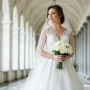 A bride wearing an elegant white lace wedding gown and veil stands in a bright arched hallway, holding a bouquet of white roses.