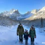 Realistic winter travel scene showing a family walking through a snowy U.S. landscape with mountains.